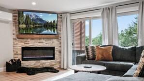 A living room with a stone fireplace, a large flat-screen TV mounted above it, a grey sofa with patterned cushions, and a view of trees and mountains through large windows.