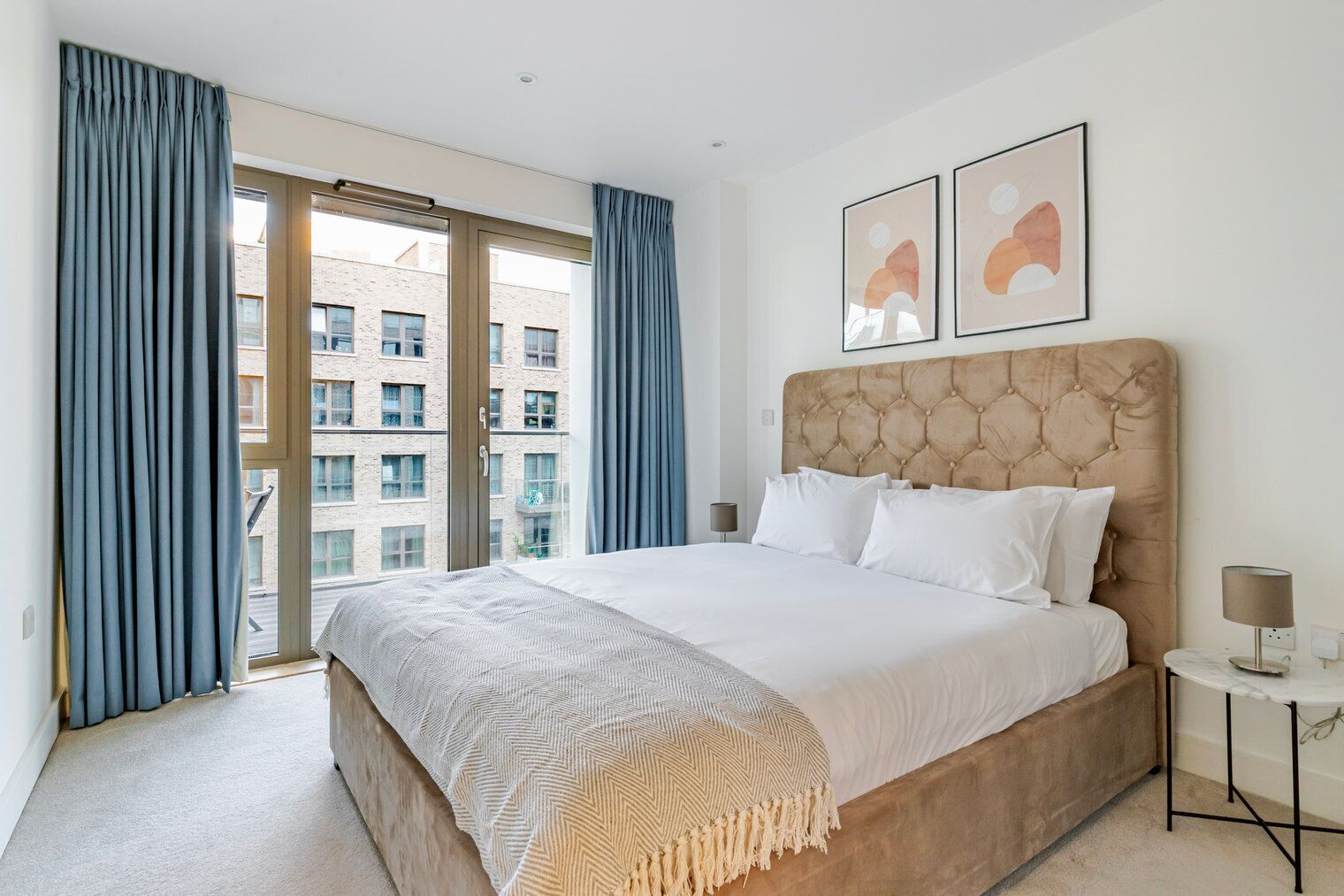 A modern hotel room with a large bed, a view of the city through floor-to-ceiling windows, and two abstract art pieces on the wall.