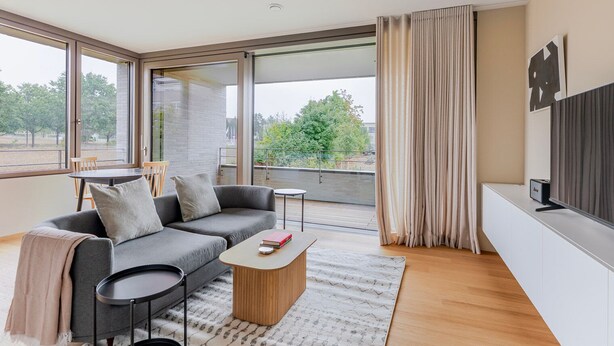 A modern living room with a grey sofa, a coffee table, and a TV on a white cabinet. There is a balcony with a view of trees and buildings. - Blueground | Kirchberg, balcony, nr Parc de Merl (Luxembourg)