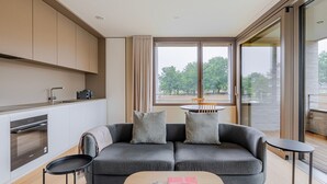 A modern living room with a grey sofa, a small round table, and a kitchen area with a built-in oven and cabinets. - Blueground | Kirchberg, balcony, nr Parc de Merl (Luxembourg)