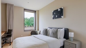 A hotel room with a bed, a desk, a chair, a nightstand, and a window with a view of trees. - Blueground | Kirchberg, balcony, nr Parc de Merl (Luxembourg)