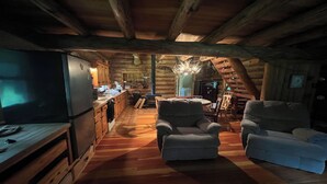 Interior de una cabaña de troncos con cocina, dos sillones, una mesa de comedor y una chimenea.