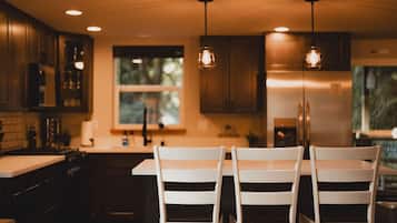 A modern kitchen with dark wooden cabinets, a central island with white chairs, pendant lighting, and a large window.