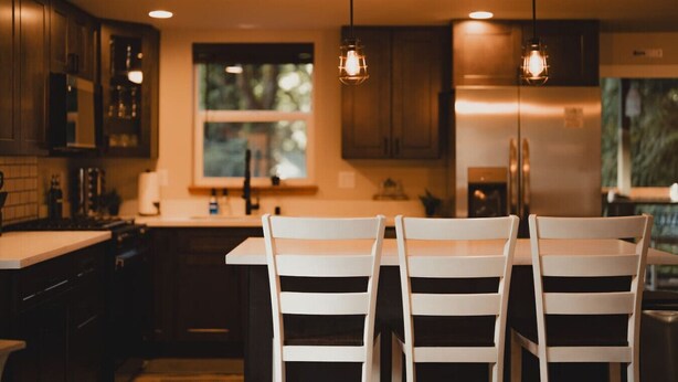Una cocina moderna con armarios de madera oscura, una isla central con sillas blancas, iluminación colgante y un ventanal grande.