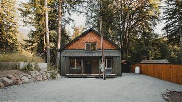 A small cabin with a sloped roof, a front porch with two chairs, and a gravel driveway.