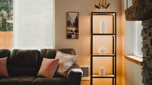 Una sala de estar acogedora con un sofá de cuero oscuro, un cuadro de paisaje enmarcado, una estantería alta de color negro con adornos blancos y una pared de piedra.