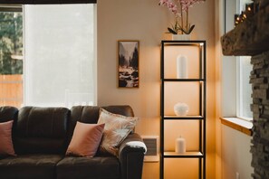 A cozy living room with a dark leather sofa, a framed landscape painting, a tall black shelving unit with white decorative items, and a stone wall.