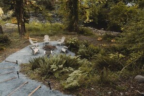 A circular seating area with white chairs around a central fire pit, surrounded by ferns and trees.