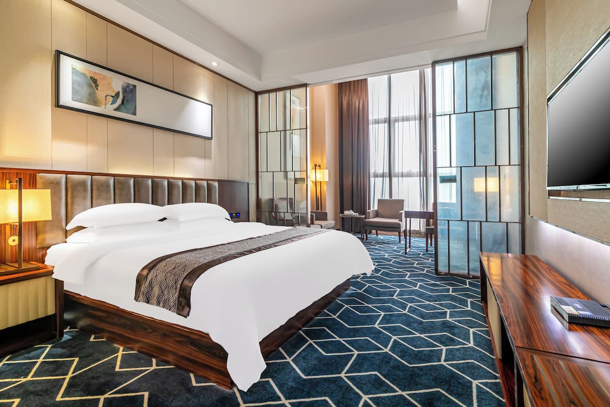 A hotel room with a large bed, a desk, a chair, a TV, and a large window with curtains.