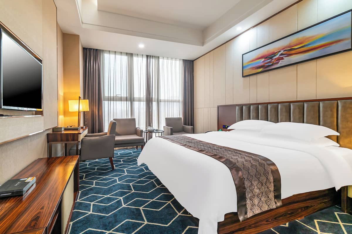 A hotel room with a large bed, a desk with a laptop, a TV, a chair, a small table, and a window with curtains.