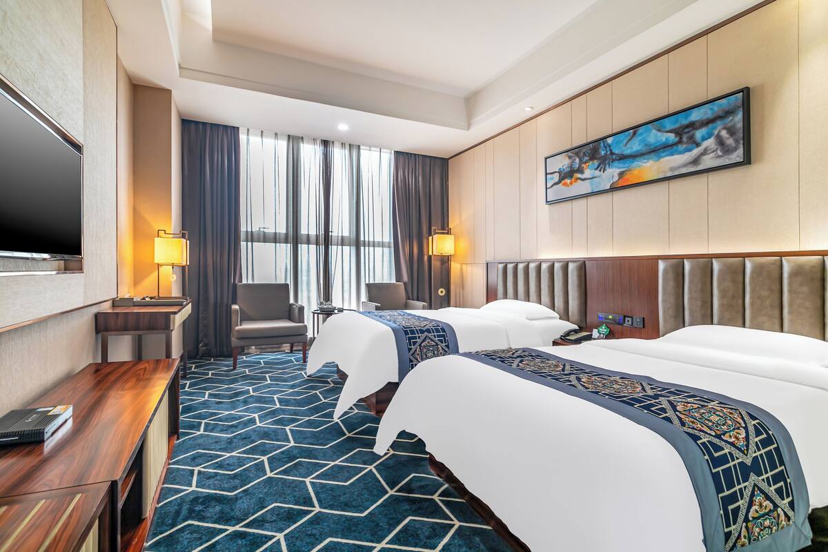 A hotel room with two beds, a desk, a TV, and a large window with curtains.