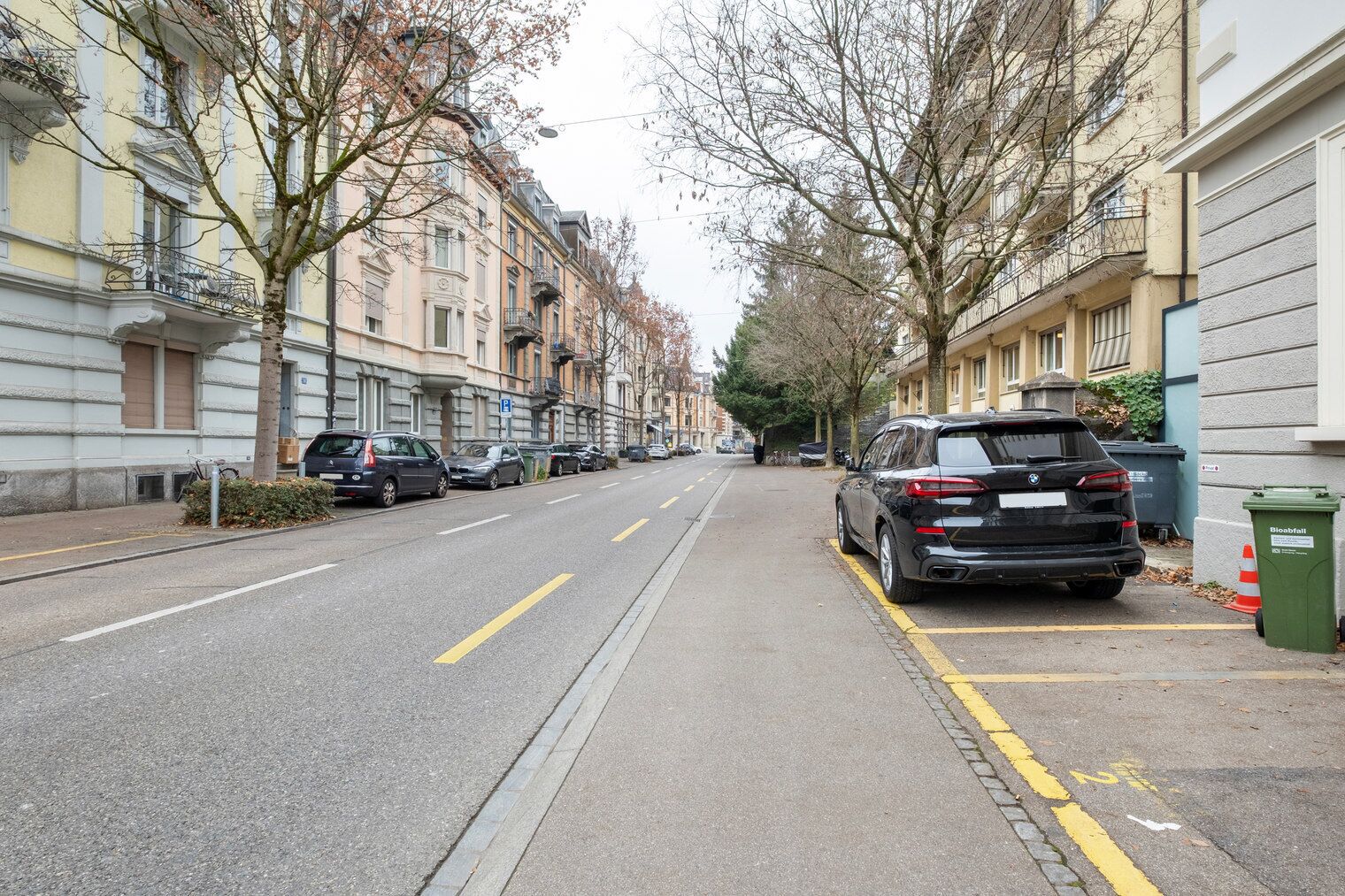 A street lined with multi-story residential buildings, parked cars, and a green bin labeled "Biofall".