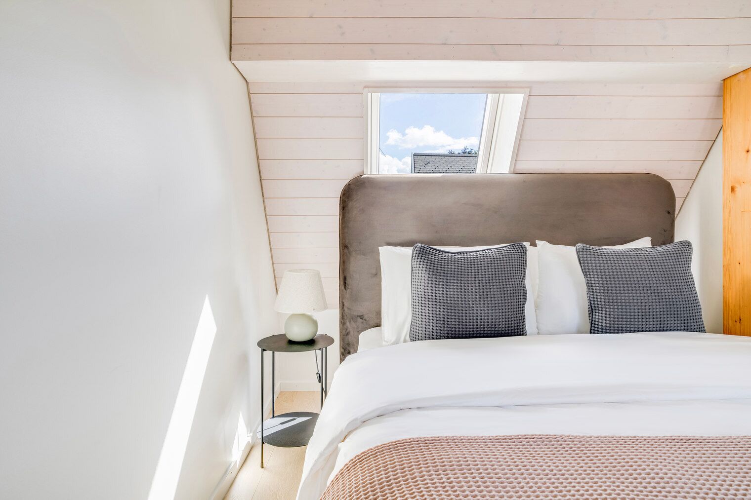 Une chambre avec un plafond en pente, un puits de lumière, un lit avec une literie blanche, deux coussins à motifs, une lampe de chevet et une petite table ronde.