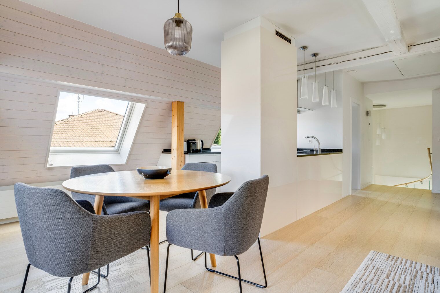 A modern dining area with a round wooden table and grey upholstered chairs, a large window, and a view of the kitchen.