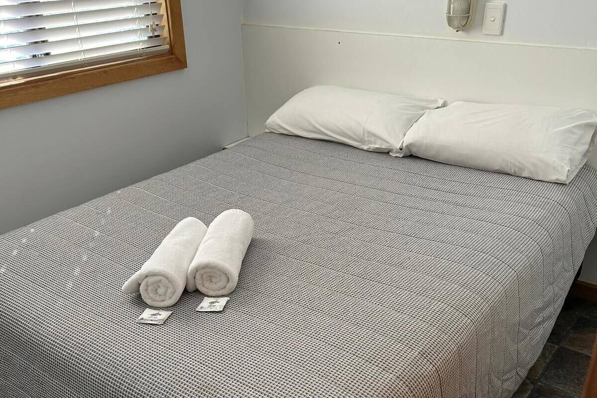 Un lit simple avec une couvre-lit à motifs, deux serviettes enroulées, un luminaire fixé au mur et un tableau encadré.