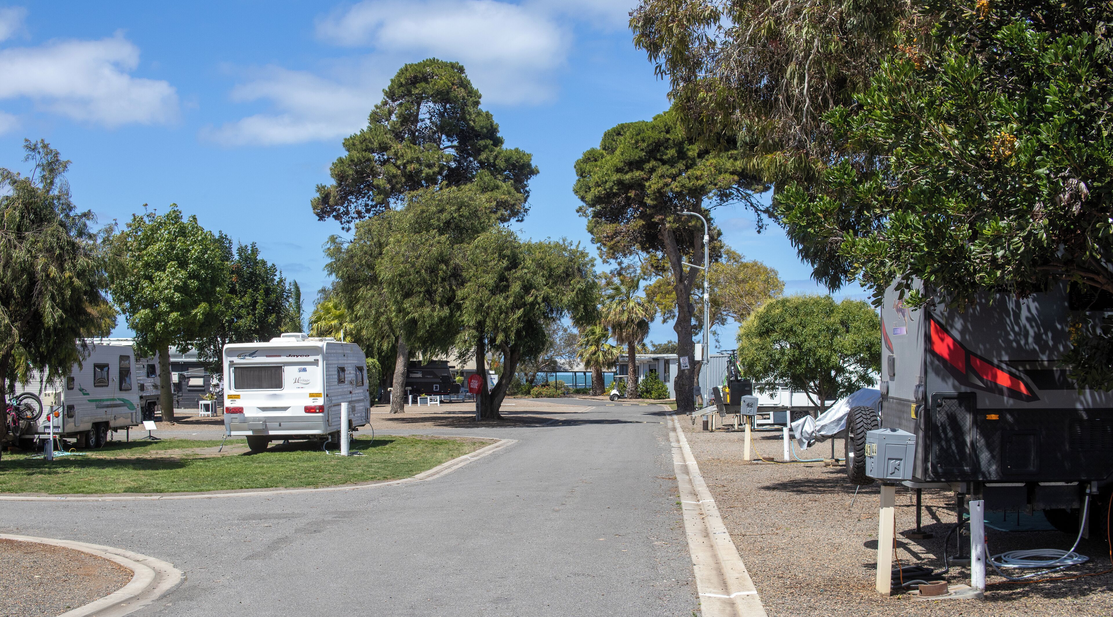 Ein Campingplatz mit Reisemobilen, die an einer befestigten Straße geparkt sind, umgeben von Bäumen und blauem Himmel.