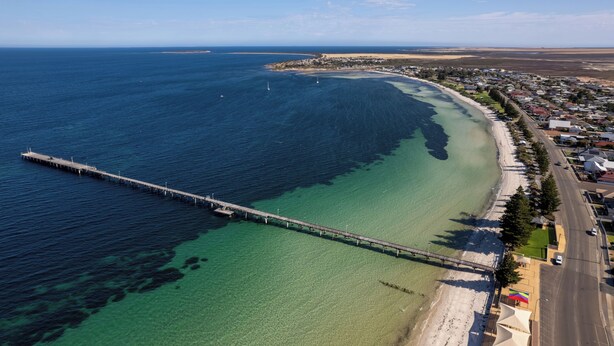 An aerial view of a coastal area with a long pier extending into the sea, a beach, and a residential area with houses and roads. - Tumby Bay Caravan Park (Tumby Bay)