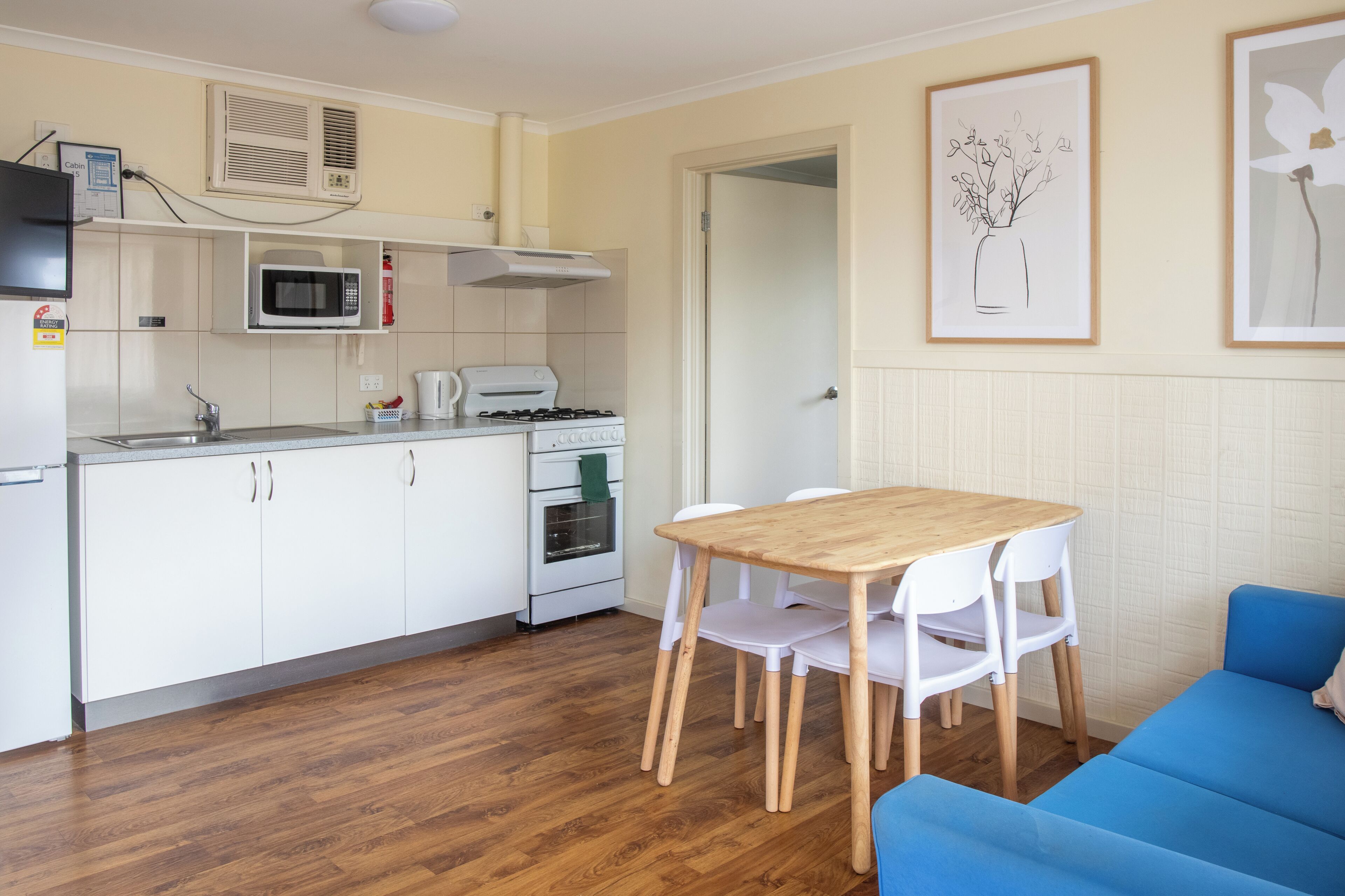 Une cuisine compacte avec des meubles blancs, un four à micro-ondes, une cuisinière et un évier. On y trouve une table à manger avec des chaises et un canapé bleu.