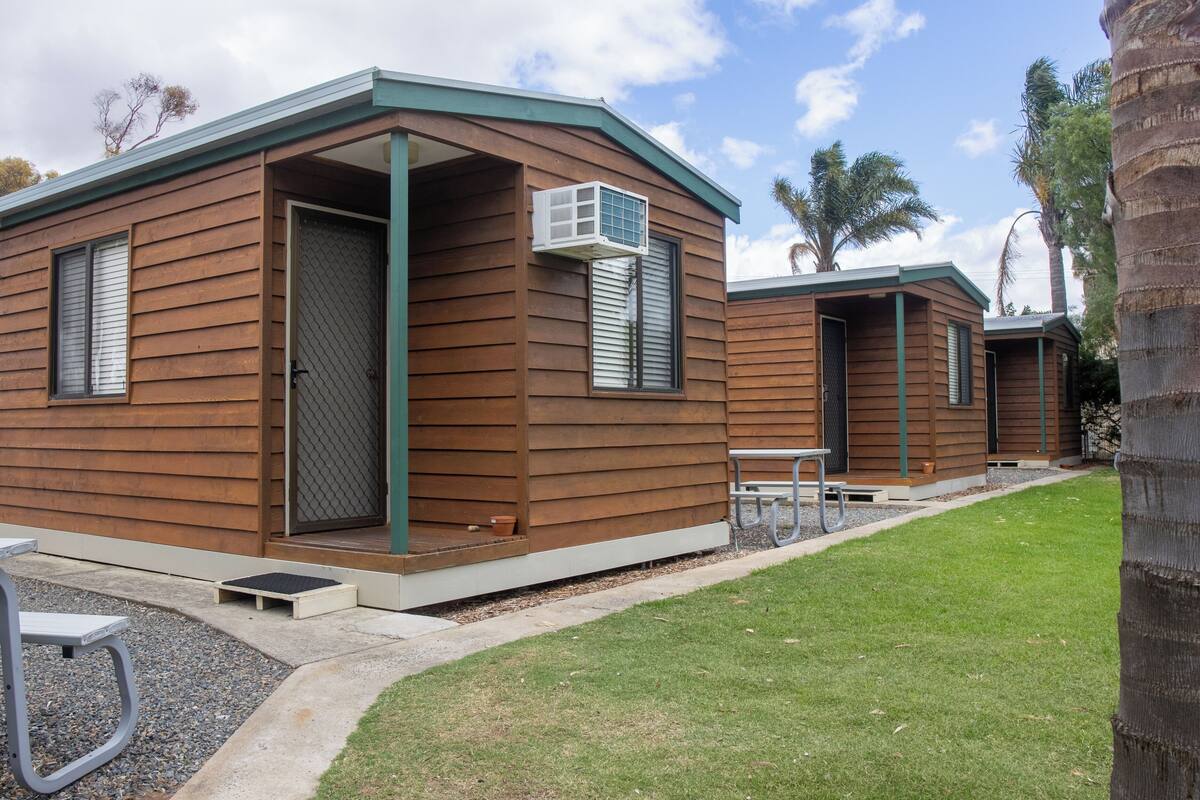 Une rangée de petites cabanes en bois, dotées d’un toit vert, chacune ayant une porte et une fenêtre, ainsi qu’un petit espace patio avec une table et des bancs.