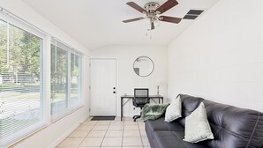 Un salon moderne avec un canapé en cuir noir, un grand miroir rond, un ventilateur de plafond et de grandes fenêtres équipées de stores.