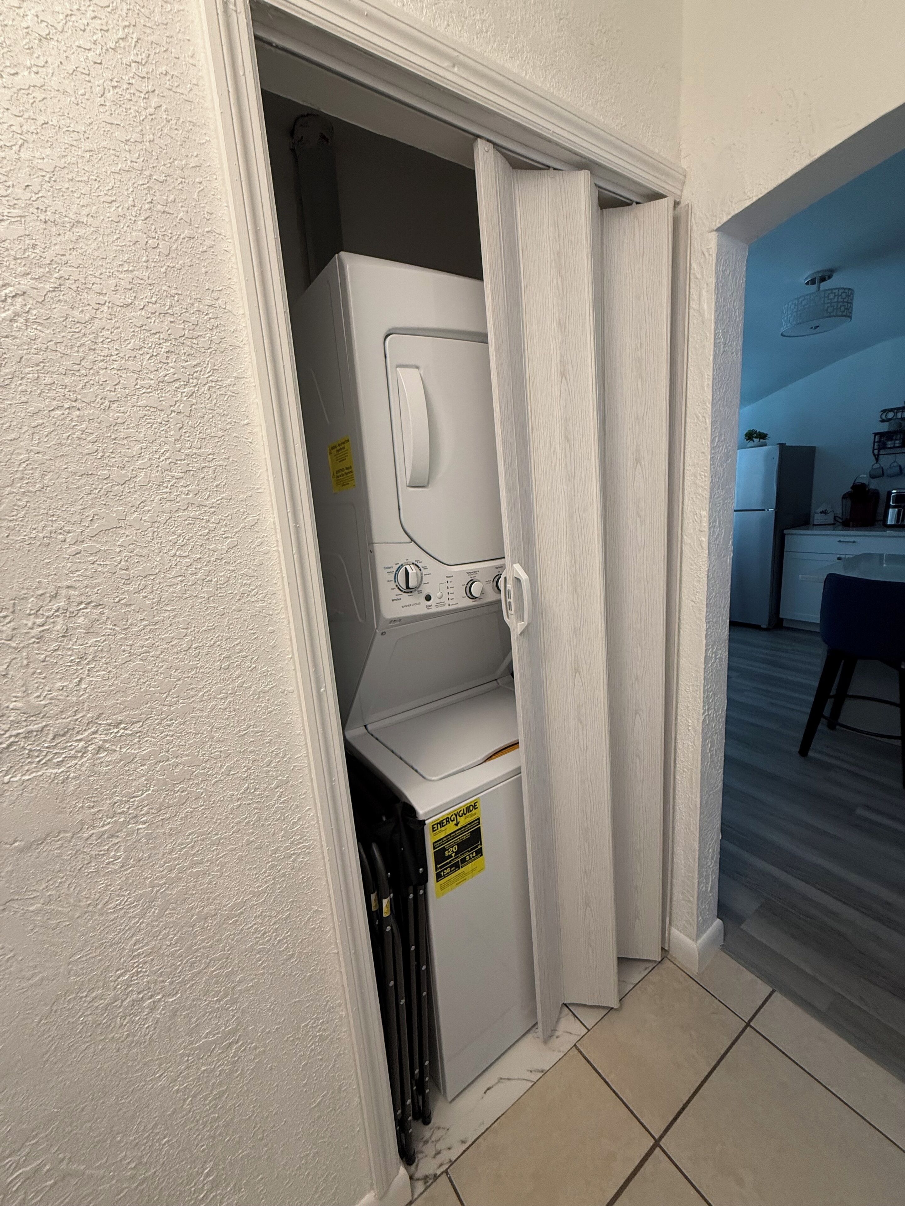 Un lavadero con lavadora y secadora, un letrero que indica eficiencia energética, y una vista parcial de una cocina con refrigerador y una planta.