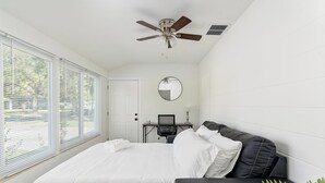 Une chambre à coucher comprenant un lit, un bureau, une chaise, un ventilateur de plafond, un miroir et une fenêtre avec des stores.