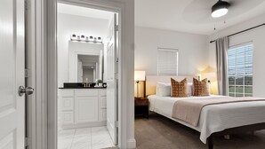 Ein Schlafzimmer mit Bett, Nachttischlampen, einem Nachttisch, einem Fenster mit Jalousien und einem durchgegangenen Türspalte sichtbaren Badezimmer.