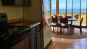 A kitchen with a stove, refrigerator, and wooden cabinets. There is a dining area with a table and chairs, and a view of the ocean through large windows.
