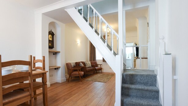 Un vaste espace de vie comprenant une table et des chaises en bois pour la salle à manger, un canapé en cuir marron et un escalier avec une rampe en verre menant à l’étage supérieur.