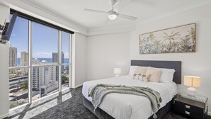 Une chambre d’hôtel avec une grande fenêtre, un lit avec du linge de maison blanc, un ventilateur de plafond et une table de chevet avec une lampe.