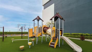 Un parque infantil con toboganes y estructuras para escalar, rodeado de un césped cuidado y un edificio al fondo.