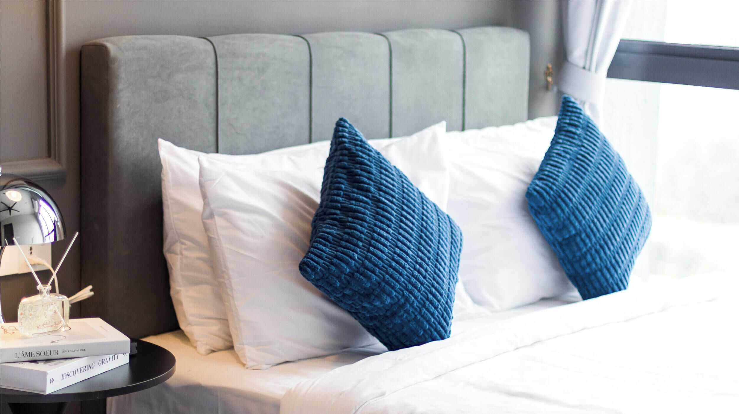 A neatly made bed with white and blue pillows, a bedside table with a lamp and books, and a grey upholstered headboard.