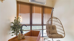 A room with a hanging chair, a wooden table with a plant and a diffuser, and a large window with blinds. - KIP Core Soho Suite by Five Senses (Sepang)