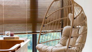 A hanging chair with a cushion in a room with wooden blinds and a view of greenery outside.