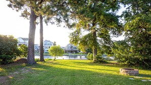 A well-maintained grassy area with a stone fire pit, surrounded by mature trees and a body of water in the background.