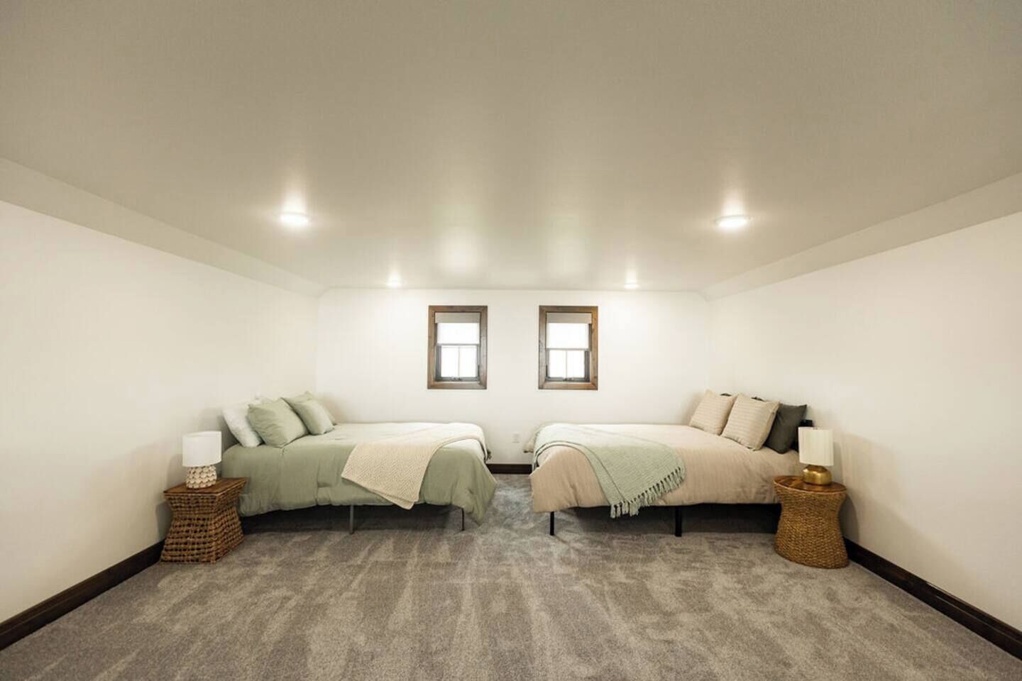 Una habitación amplia con dos camas, cada una con una mesita de noche y lámpara, y dos ventanas.