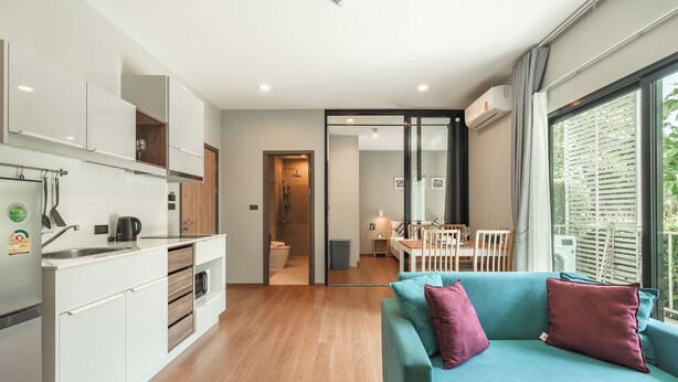 A modern kitchen with white cabinets, a built-in oven, and a coffee maker. There is a teal sofa with cushions in the living area, and a dining table with chairs. - 1BR Pool Access Near Rawai Beach C222 (Rawai)