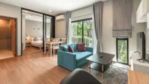 A modern living room with a sofa, a dining table, and a TV. There is a sliding glass door leading to a balcony with a view of greenery. - 1BR Pool Access Near Rawai Beach C222 (Rawai)