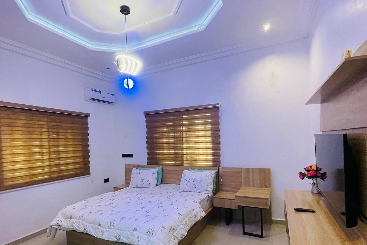 Ein modernes Schlafzimmer mit sechseckigem Deckenstil, einem Bett mit floraler Bettwäsche, einem hölzernen Kopfteil, Nachttischen, einem Fernseher und einer kleinen Vase mit Blumen.