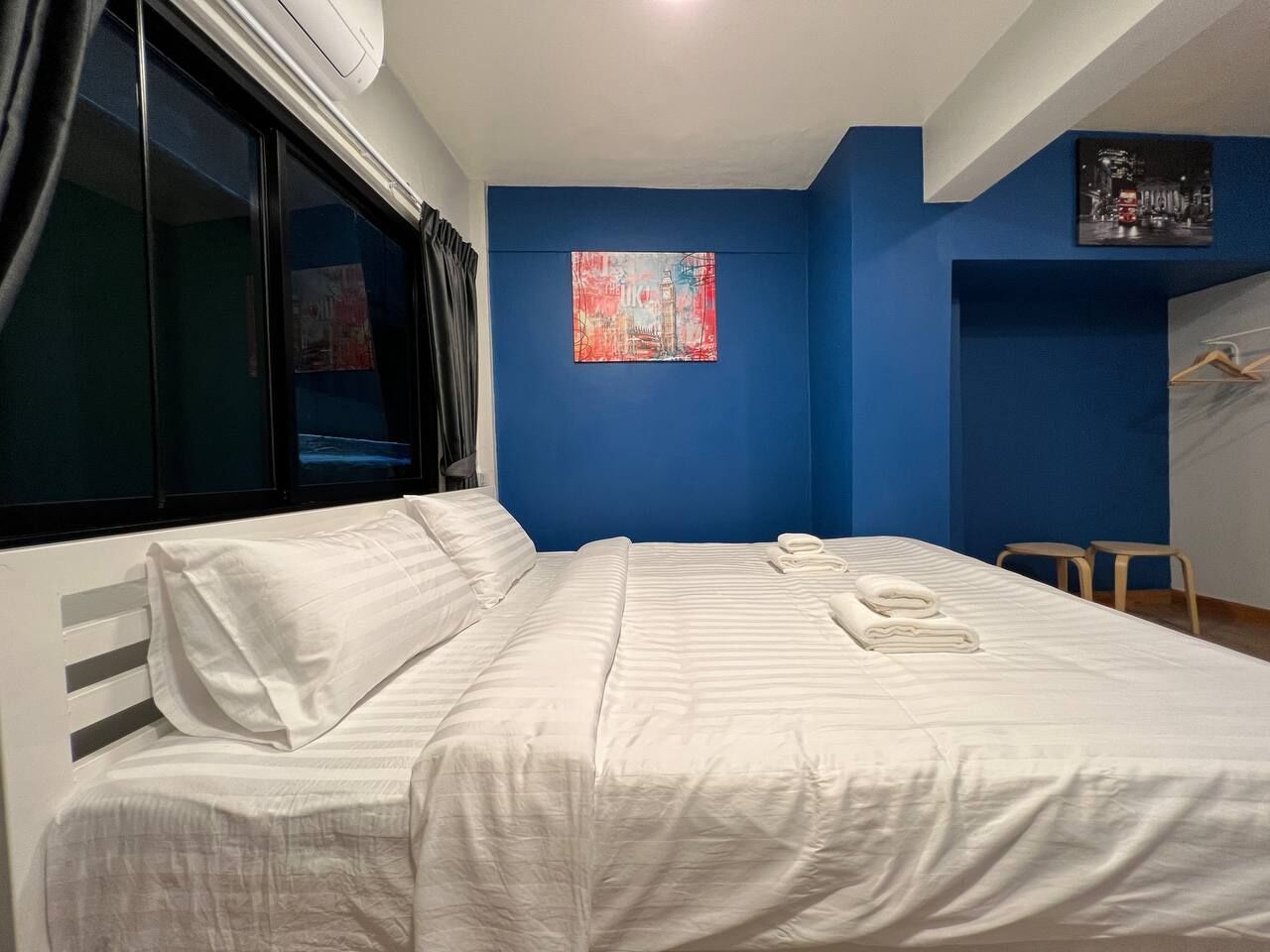 A hotel room with a bed, a window, a blue accent wall, and a painting.