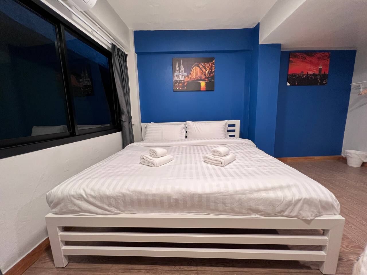 Un lit bien fait, avec du linge de lit et des serviettes blanches, une fenêtre avec des rideaux et un mur bleu orné de deux tableaux encadrés.