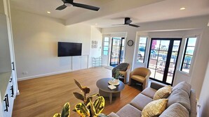 A spacious living room with a large sofa, a coffee table, and a television. There are ceiling fans, a window with a view, and a door leading to an outdoor area.