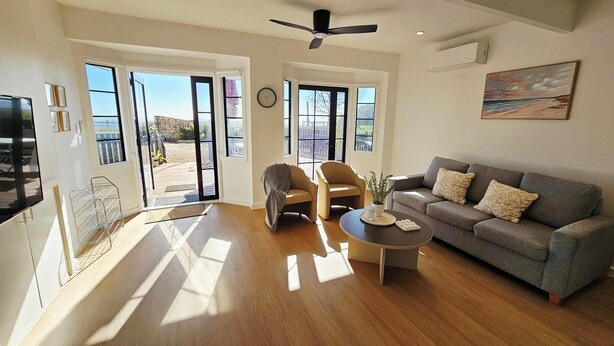 A spacious living room with a grey sofa, a coffee table, and two armchairs. There are large windows providing natural light and a view of the outdoors.