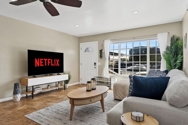 Un salon moderne avec un téléviseur à écran plat affichant le logo « NETFLIX », un canapé, une table basse et une grande fenêtre donnant sur un quartier résidentiel.