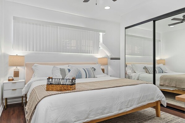 A modern bedroom with a large bed, bedside tables, a mirror, and a window with blinds.