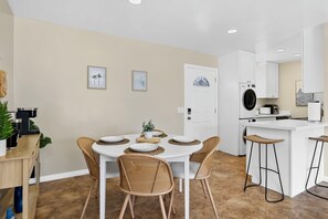 Une cuisine moderne avec des meubles blancs, un lave-linge encastré et un coin repas agrémenté de chaises en osier et d’une table blanche.