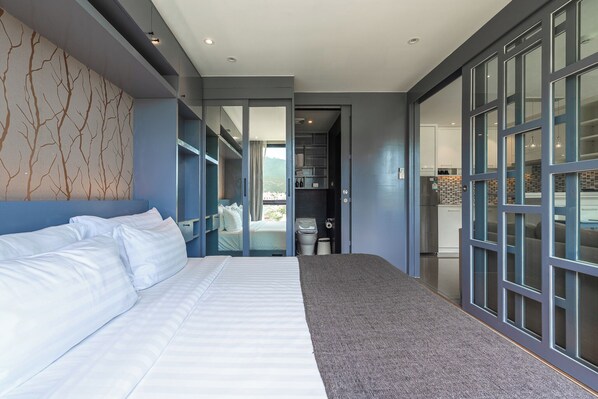 A modern hotel room with a large bed, a view through a glass door to a balcony, and a kitchen area visible through the glass wall. - 1BR Suite with Rooftop Pool C223 (Kamala)
