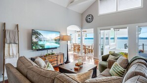 Een ruime woonkamer met een grote flatscreen televisie, comfortabele zitplaatsen, een houten salontafel en uitzicht op het landschap door grote ramen.