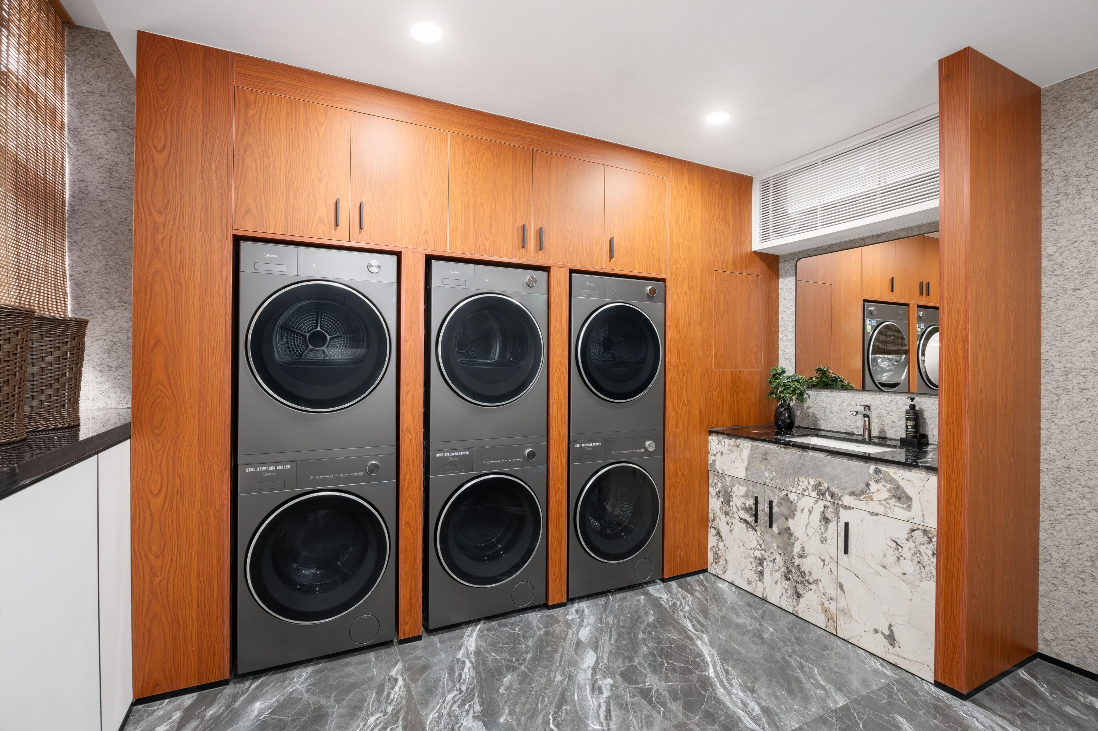 A laundry room with four front-loading washing machines, wooden cabinetry, a marble countertop, and a wall-mounted mirror.