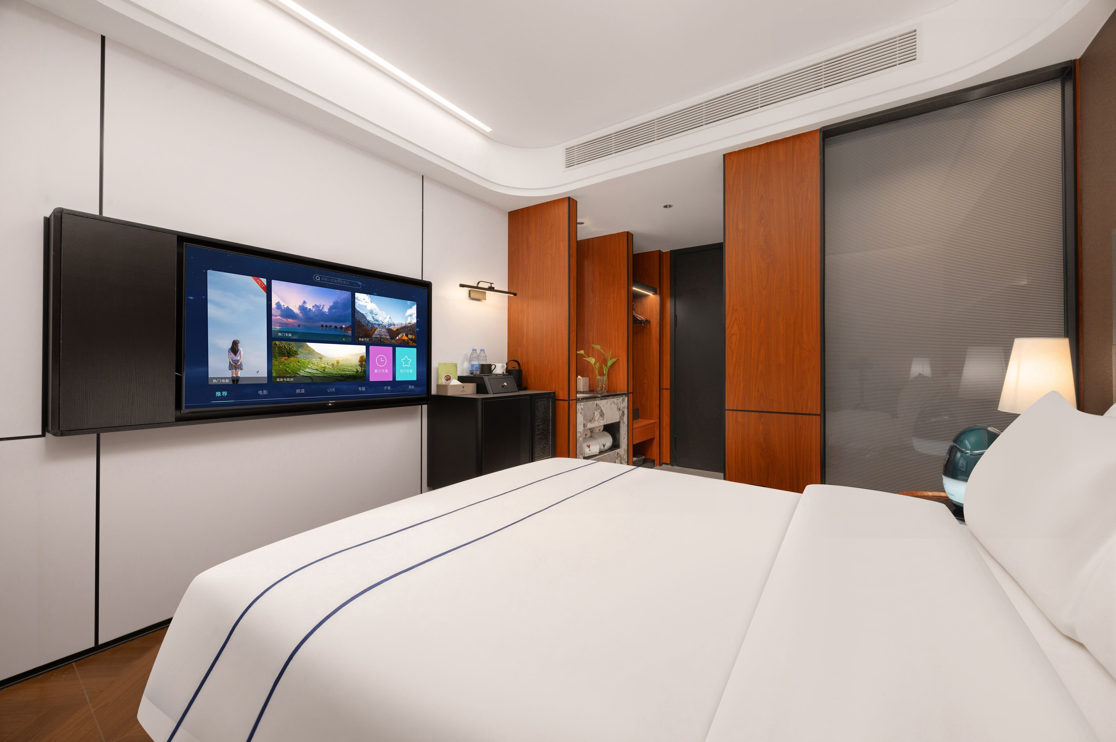 A modern hotel room with a large bed, a flat-screen TV mounted on the wall, a desk with a lamp, and a wardrobe.